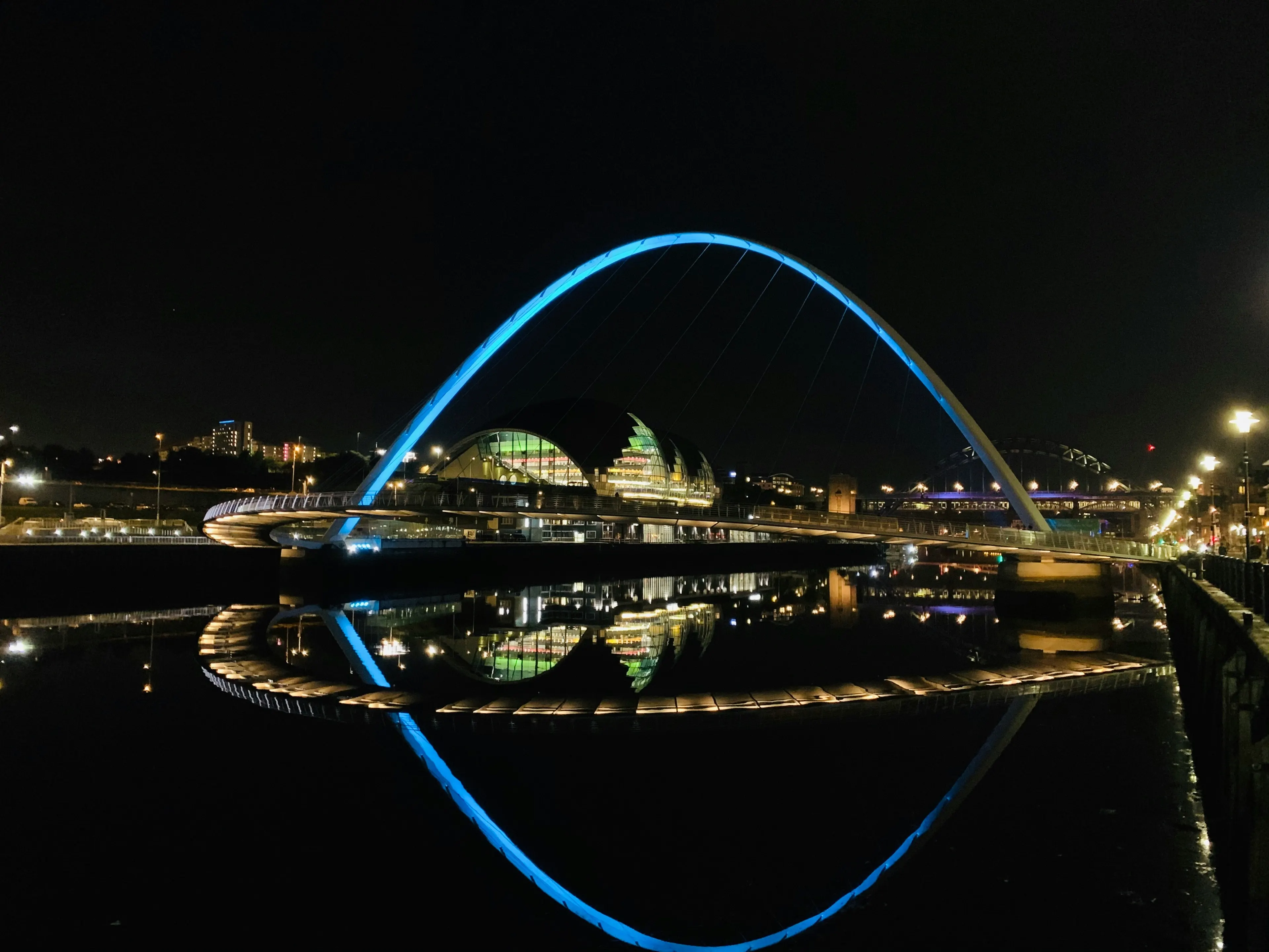 The Millennium Bridge lit cyan at night, mirrored in the River Tyne, with the Sage Gateshead behind